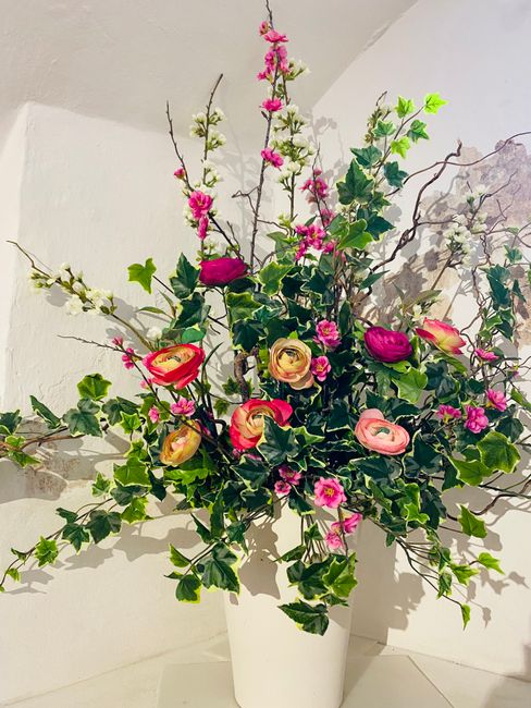 A large floral arrangement featuring a variety of fresh flowers in a white vase, including deep pink tulips, peach and pink ranunculus, and small pink blossom branches. The bouquet is lush and full, w