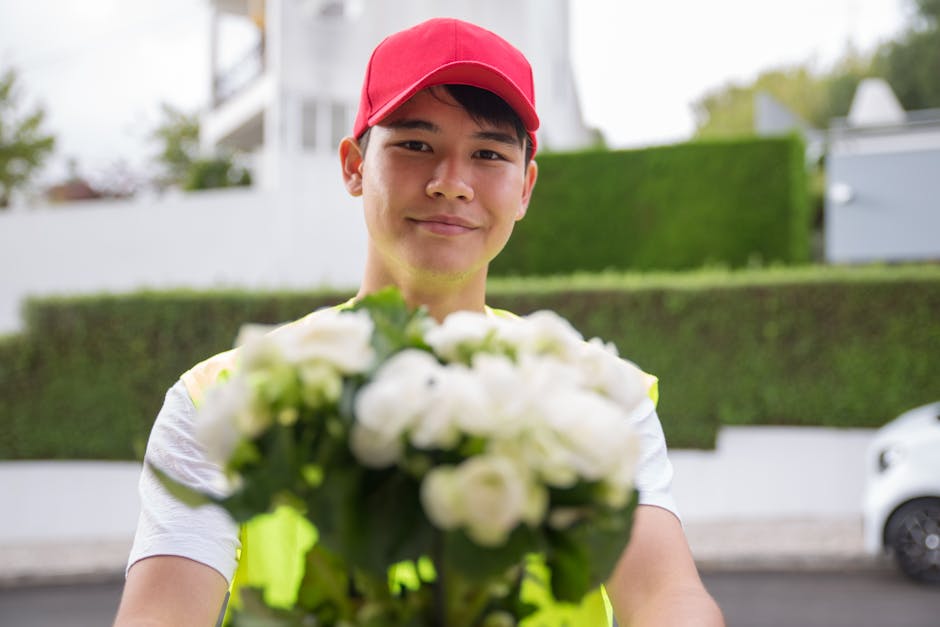 A young male flower delivery courier wearing a red cap and a neon yellow vest is smiling and holding a lush bouquet of white roses and green foliage, extending it towards the camera. The bouquet is wr