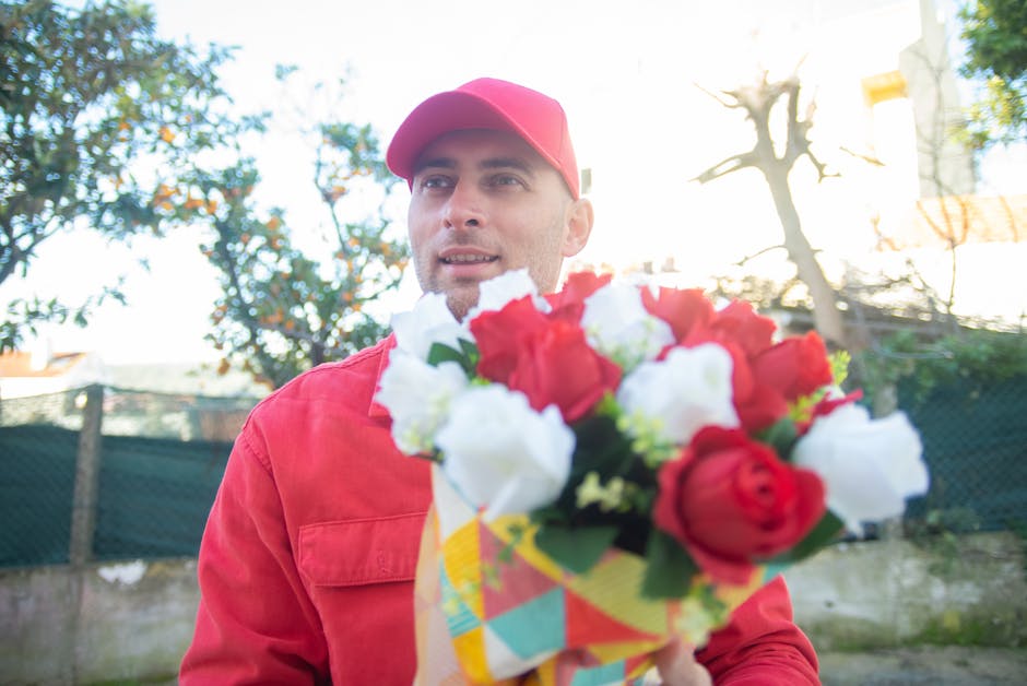A male florist wearing a red cap and matching red apron is holding a vibrant floral bouquet in front of him outdoors during daylight. The bouquet consists of large red roses, white blooms, and green f