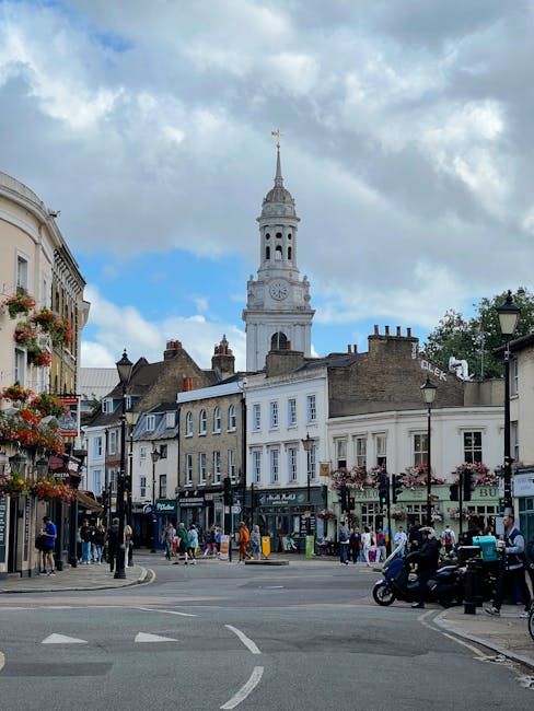 A busy town square in Chislehurst with a prominent white clock tower featuring a gold weather vane at the top, surrounded by historic buildings with shopfronts and hanging flower baskets. The scene in