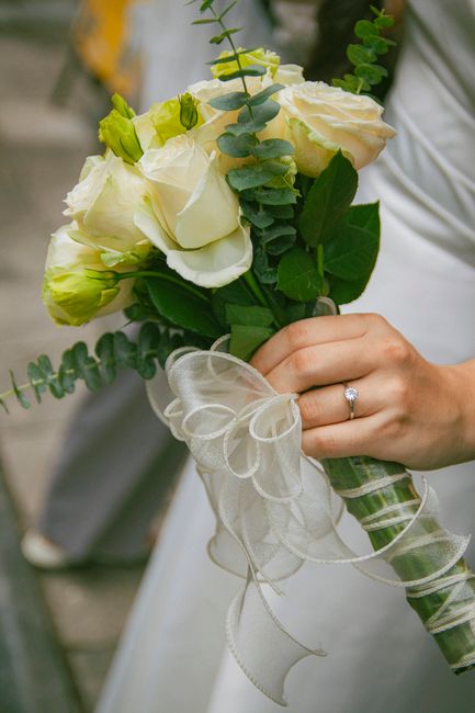 A close-up of a bride's hand holding a bridal bouquet consisting of cream and pale yellow roses, accented with sprigs of eucalyptus and greenery. The bouquet is wrapped with a delicate white ribbon ti
