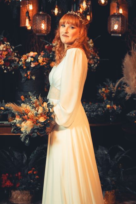 A woman with wavy red hair styled with a delicate floral headpiece is holding a floral bouquet featuring peach and blush roses, dusty miller, and dried grasses. She is wearing an elegant cream-colored